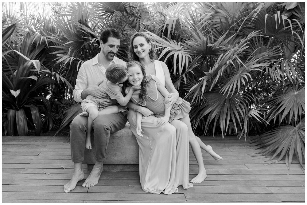 Black and white family portrait of parents with two children sitting together outdoors among tropical palm foliage.