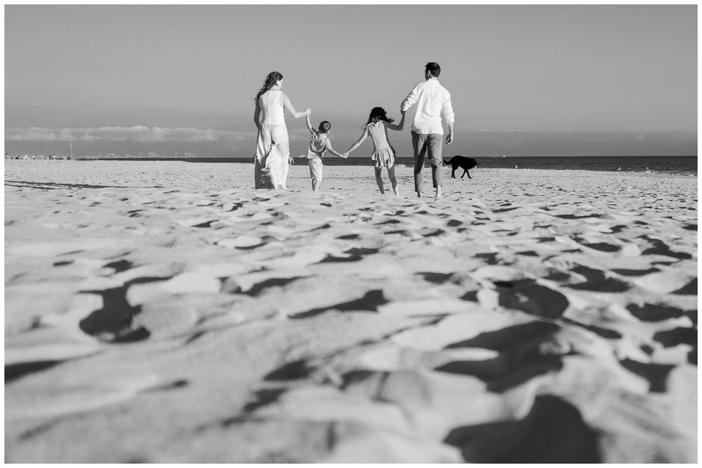 Black and white photo of a family holding hands walking on a sandy beach with a dog.