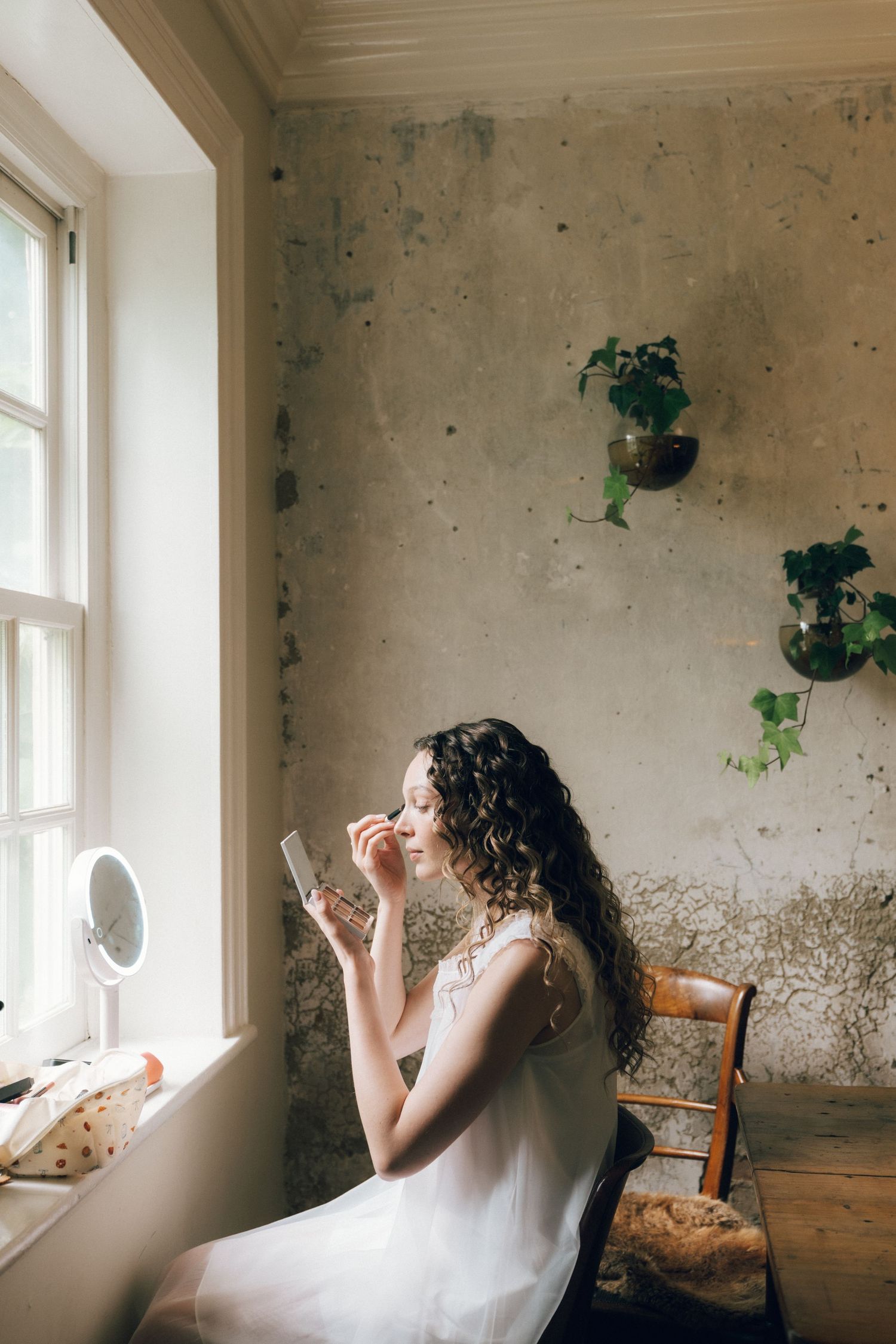 Young woman with curly hair in white dress applying makeup by window in rustic room with wall-mounted plants.
