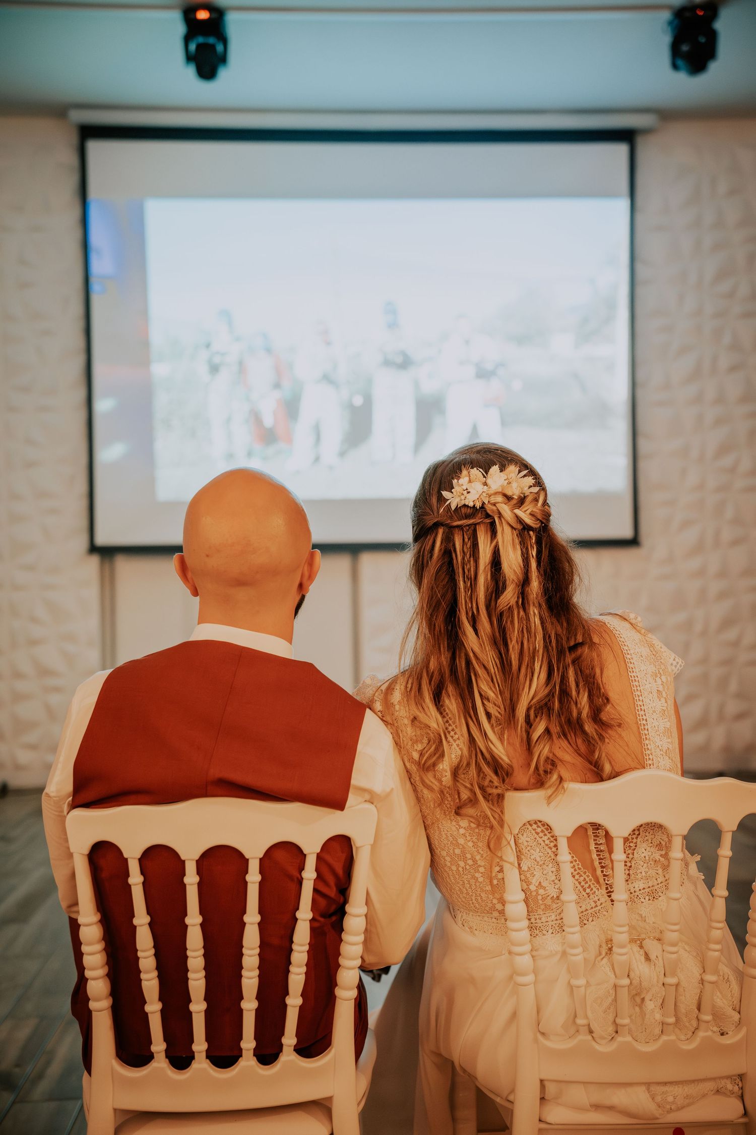Bride and groom sit in white chairs viewing wedding slideshow projected on screen during reception.