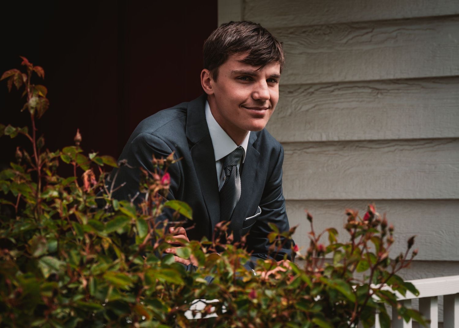 Young man in dark suit and tie smiling outdoors among lush green and red foliage near a wooden building for his Senior photos.