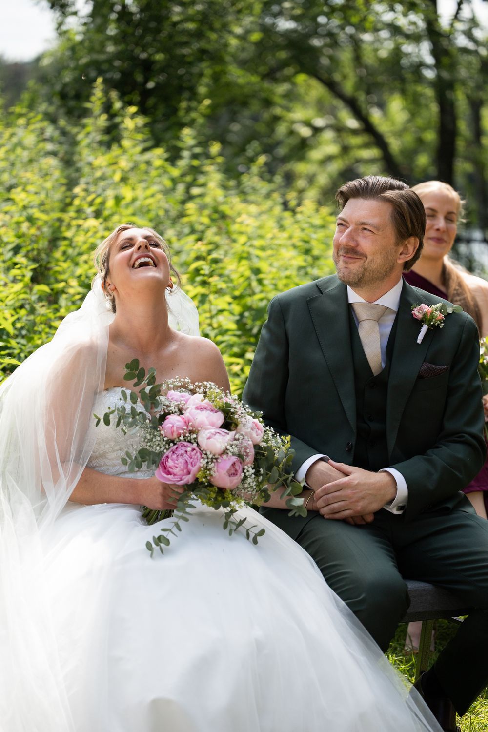 Joyful bride laughing with bouquet of pink roses beside smiling groom in green suit at outdoor wedding ceremony.