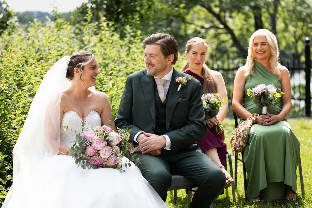 Bride and groom smiling during outdoor wedding ceremony with bridesmaids in green dresses seated behind them.