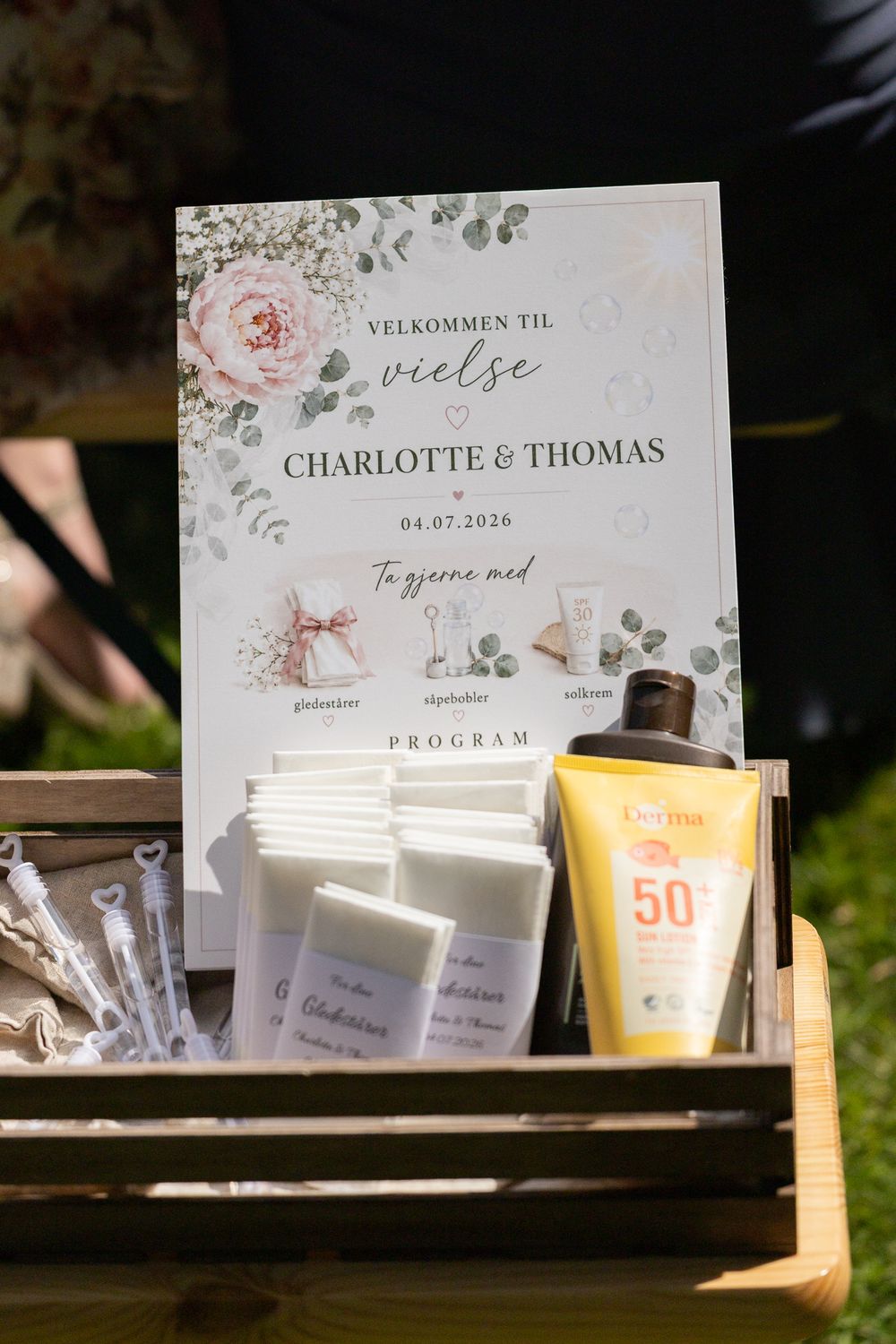Wedding welcome sign for Charlotte & Thomas dated 04.07.2026, displayed with sunscreen and program cards in a wooden crate.