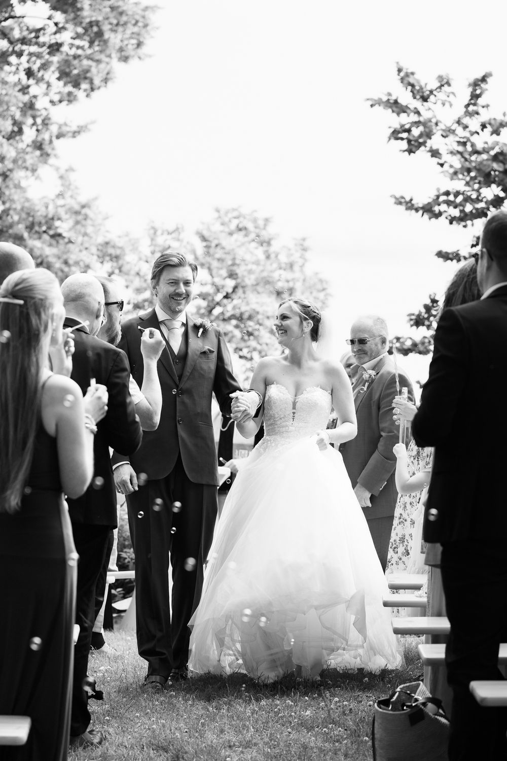 Black and white photo of a bride and groom walking down the aisle surrounded by guests blowing bubbles at outdoor wedding.
