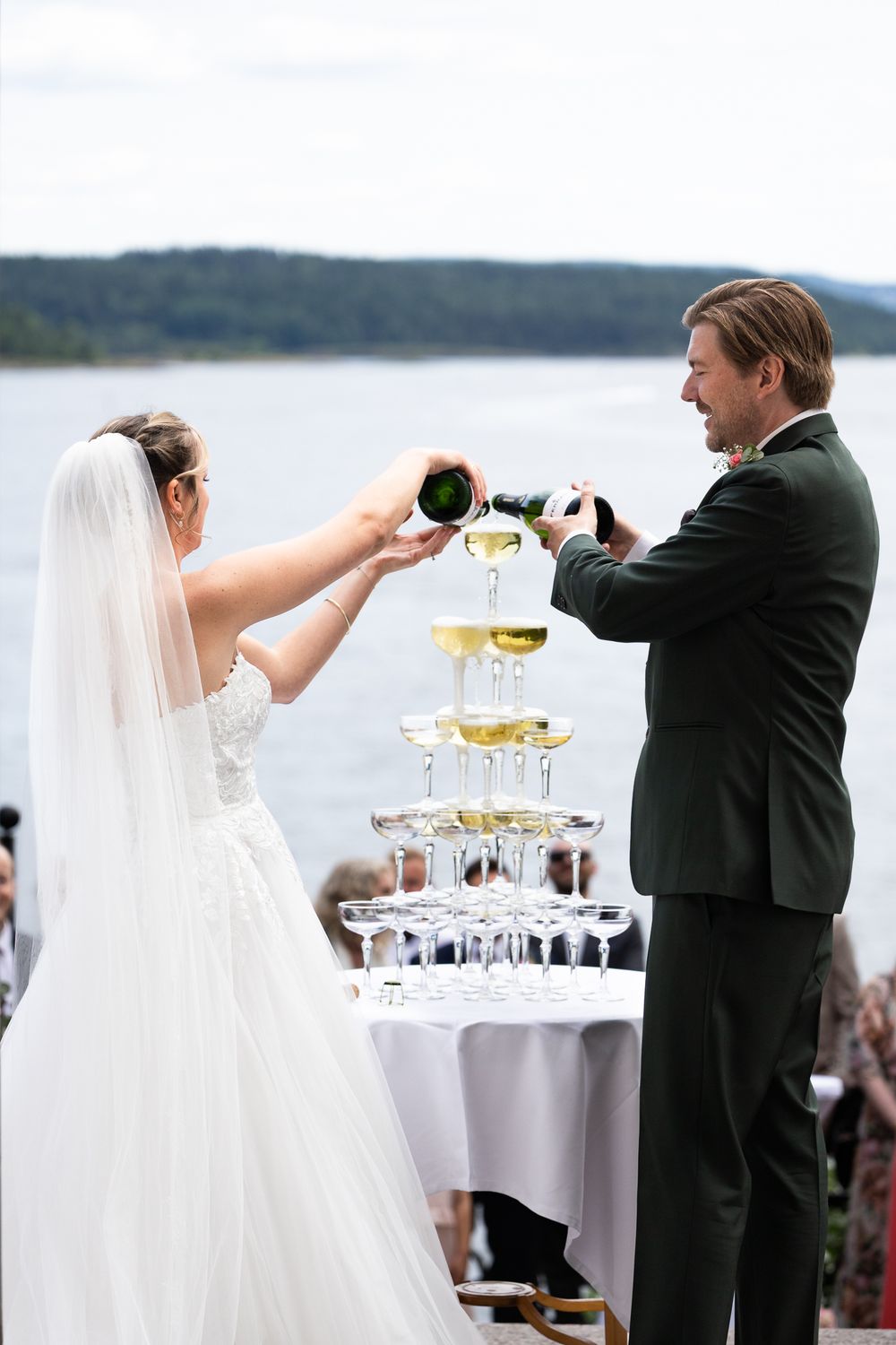 Bride and groom pouring champagne tower at outdoor waterfront wedding reception.