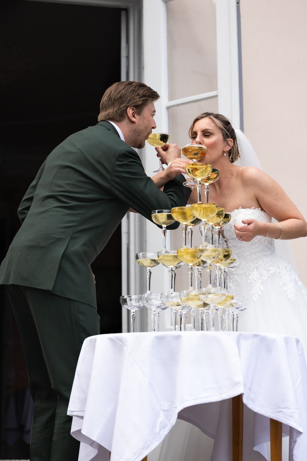 Bride and groom toasting champagne at a wedding reception beside a pyramid of champagne glasses on a white draped table.