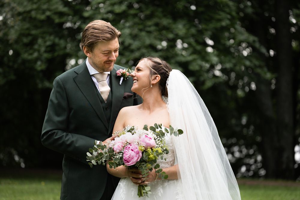 Bride and groom smiling at each other outdoors, bride holding pink peony bouquet in elegant wedding attire.
