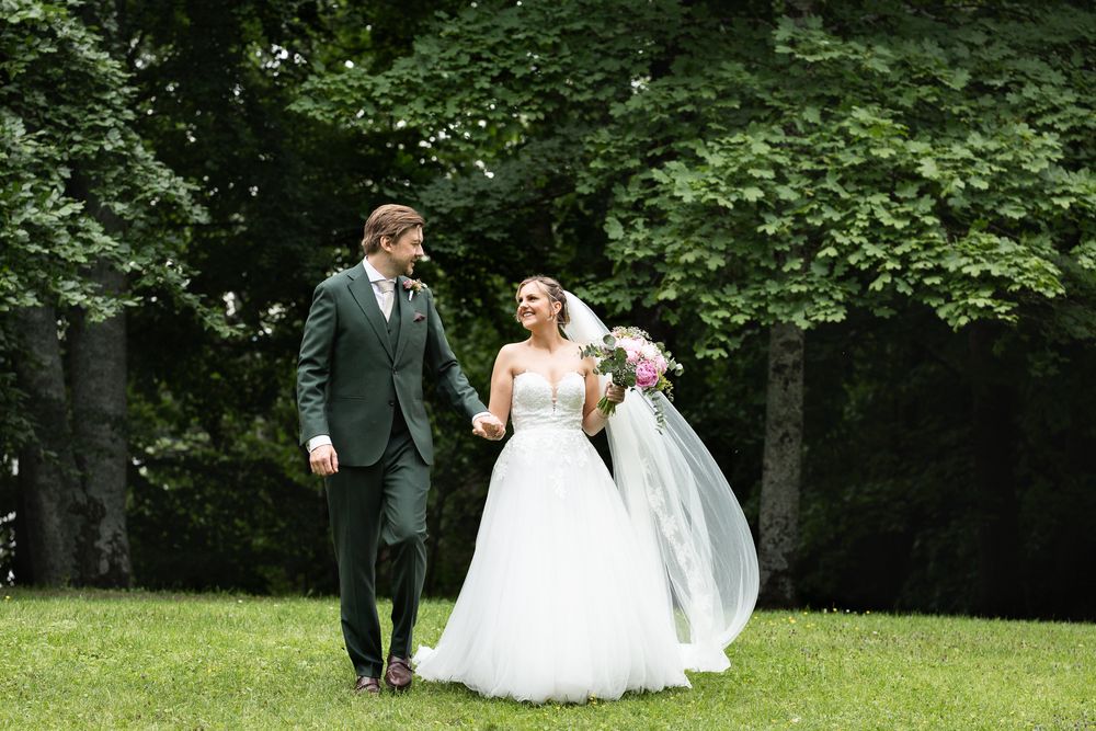 A bride and groom walk hand-in-hand on a lush green lawn, smiling at each other on their wedding day.
