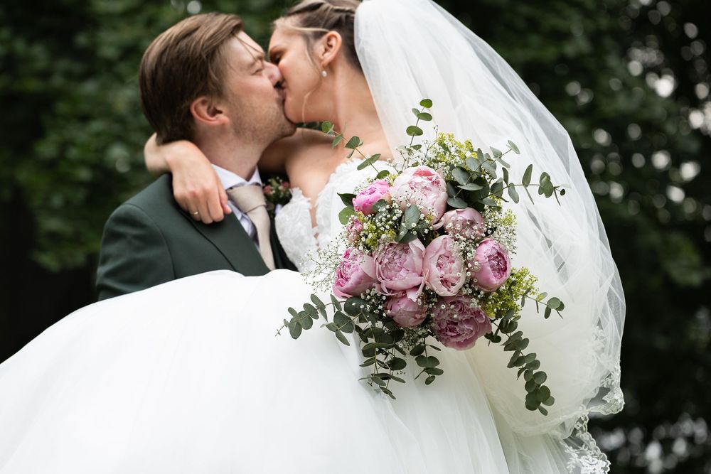 Groom lifts bride in white gown as they kiss, she holds pink rose bouquet with greenery at outdoor wedding.