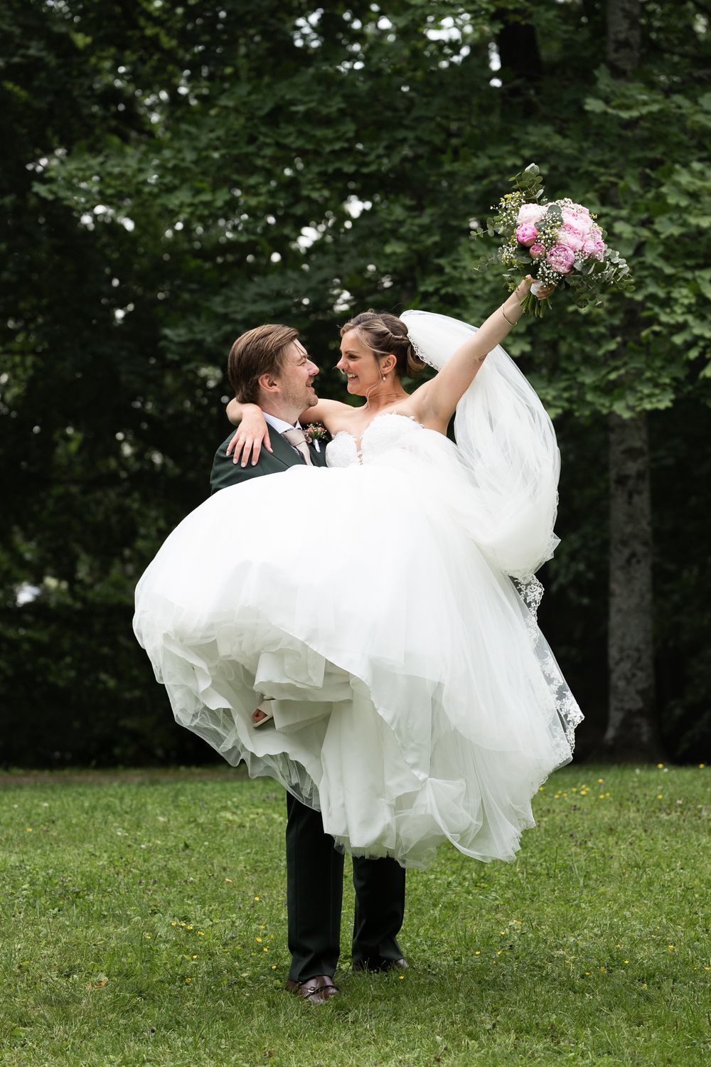 Groom lifting smiling bride in white gown outdoors, she holds pink bouquet, green trees in background.