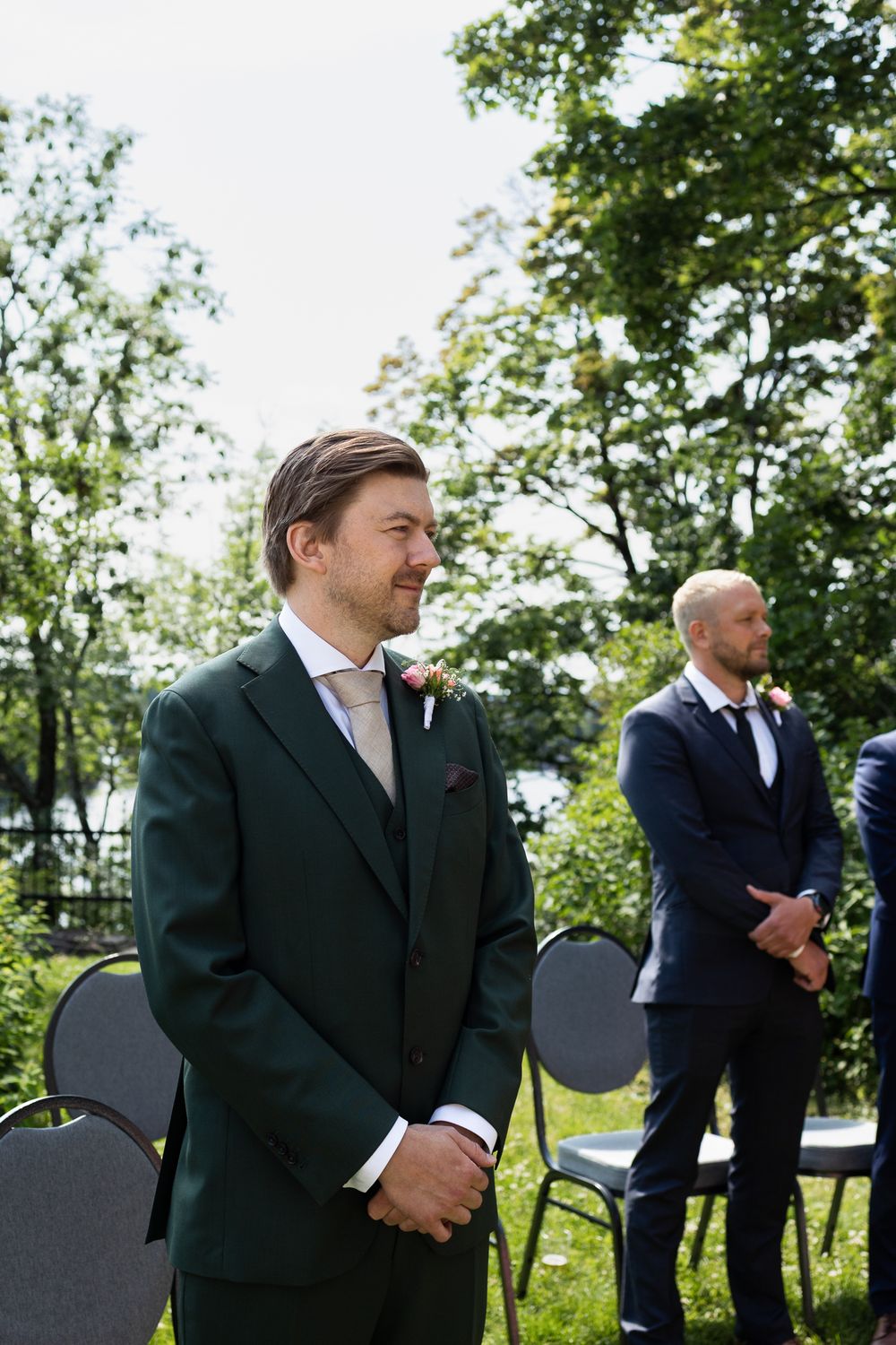 Groom in green suit smiling at outdoor wedding ceremony with groomsman in navy suit standing behind.