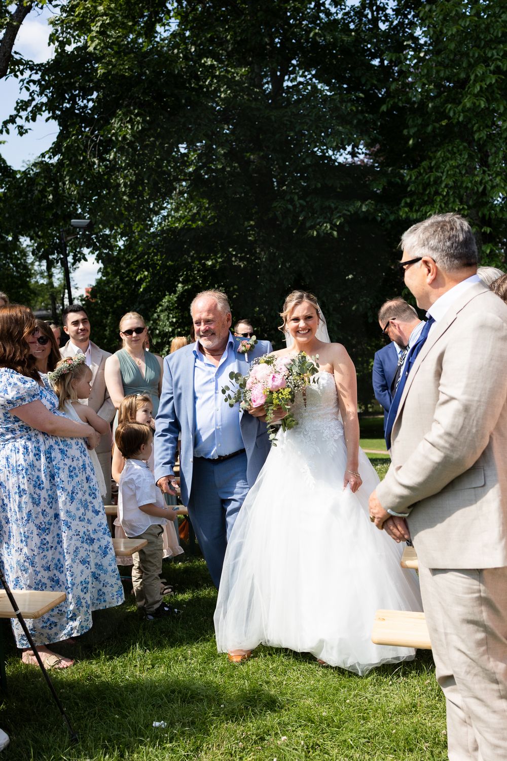 Bride in white gown walks down outdoor aisle with escort, holding pink bouquet, guests smiling in sunny garden setting.
