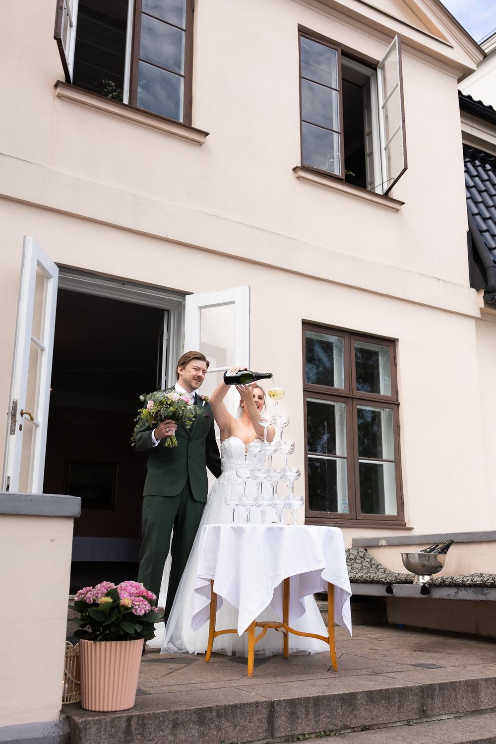 Bride and groom pouring champagne tower outside elegant venue, groom in green suit holding bouquet.