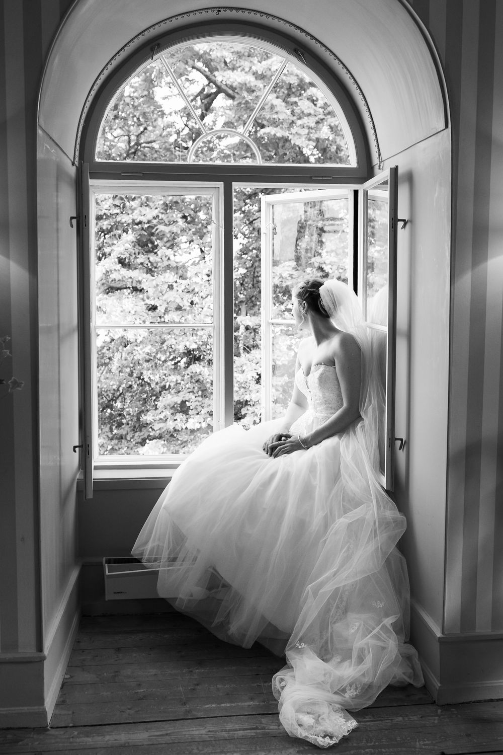 Black and white photo of a bride in a flowing gown sitting by an arched window with lush trees outside.