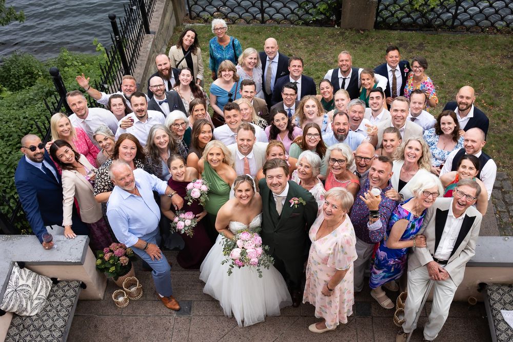 Large wedding group photo outdoors with bride and groom surrounded by smiling guests near waterfront.