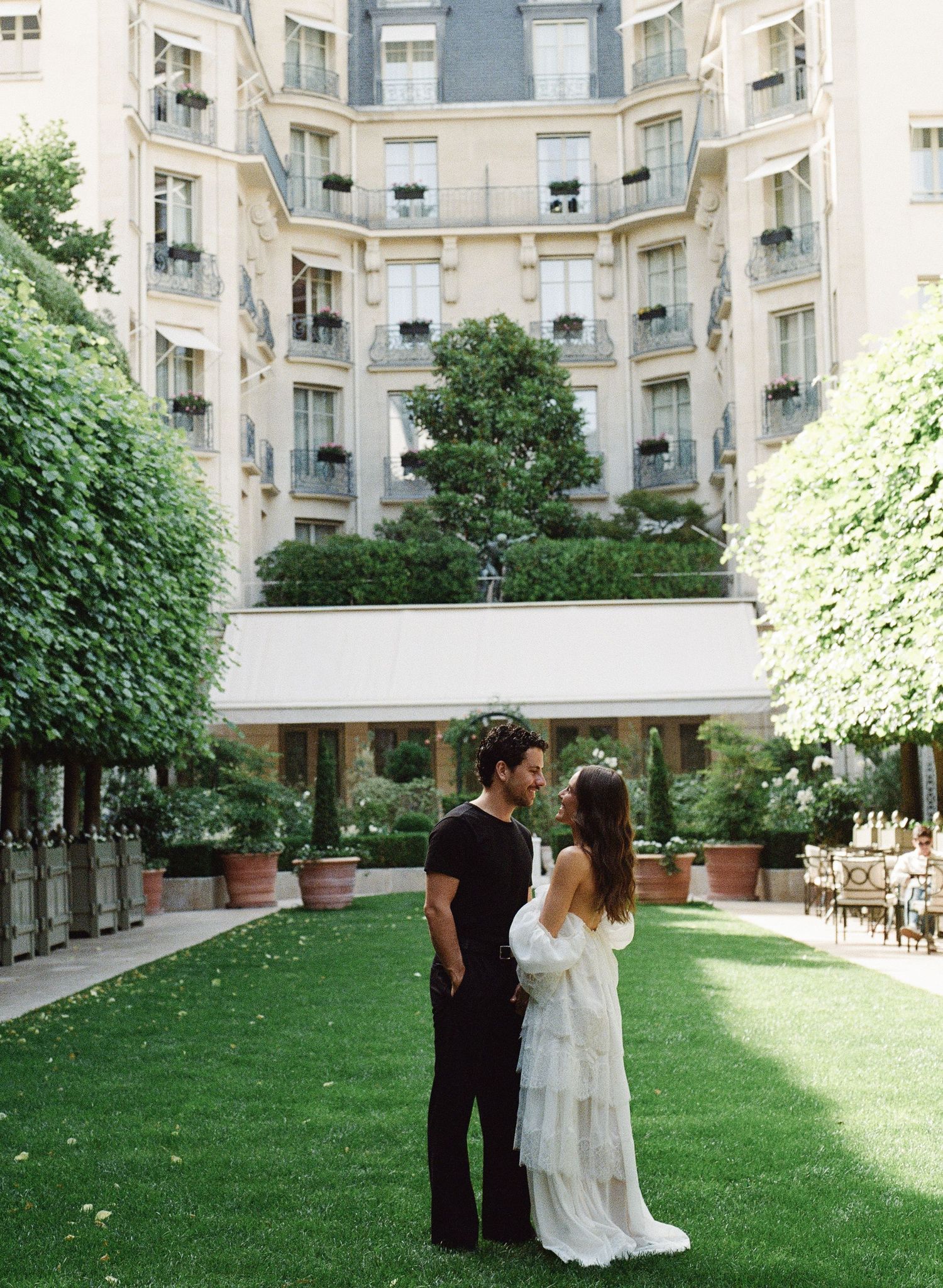 Couple standing by a stone fountain in a lush garden courtyard, woman in white dress, man in black outfit.