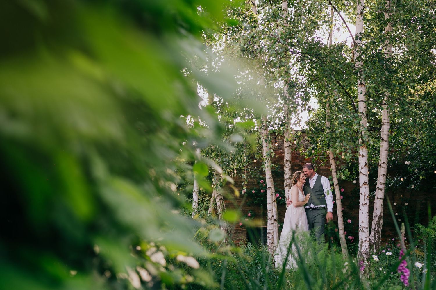 Couple embracing among birch trees in a lush green garden setting during an outdoor wedding ceremony.