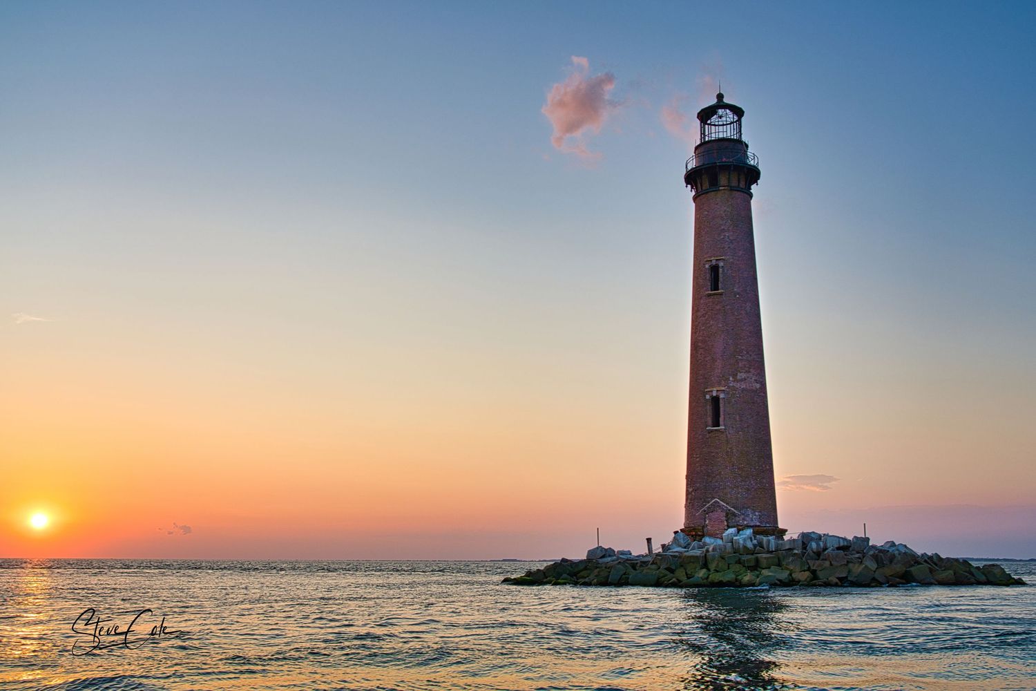 A tall, weathered brick lighthouse stands on a rocky island as the sun sets over the ocean, casting warm colors, by Steve Cole Photography