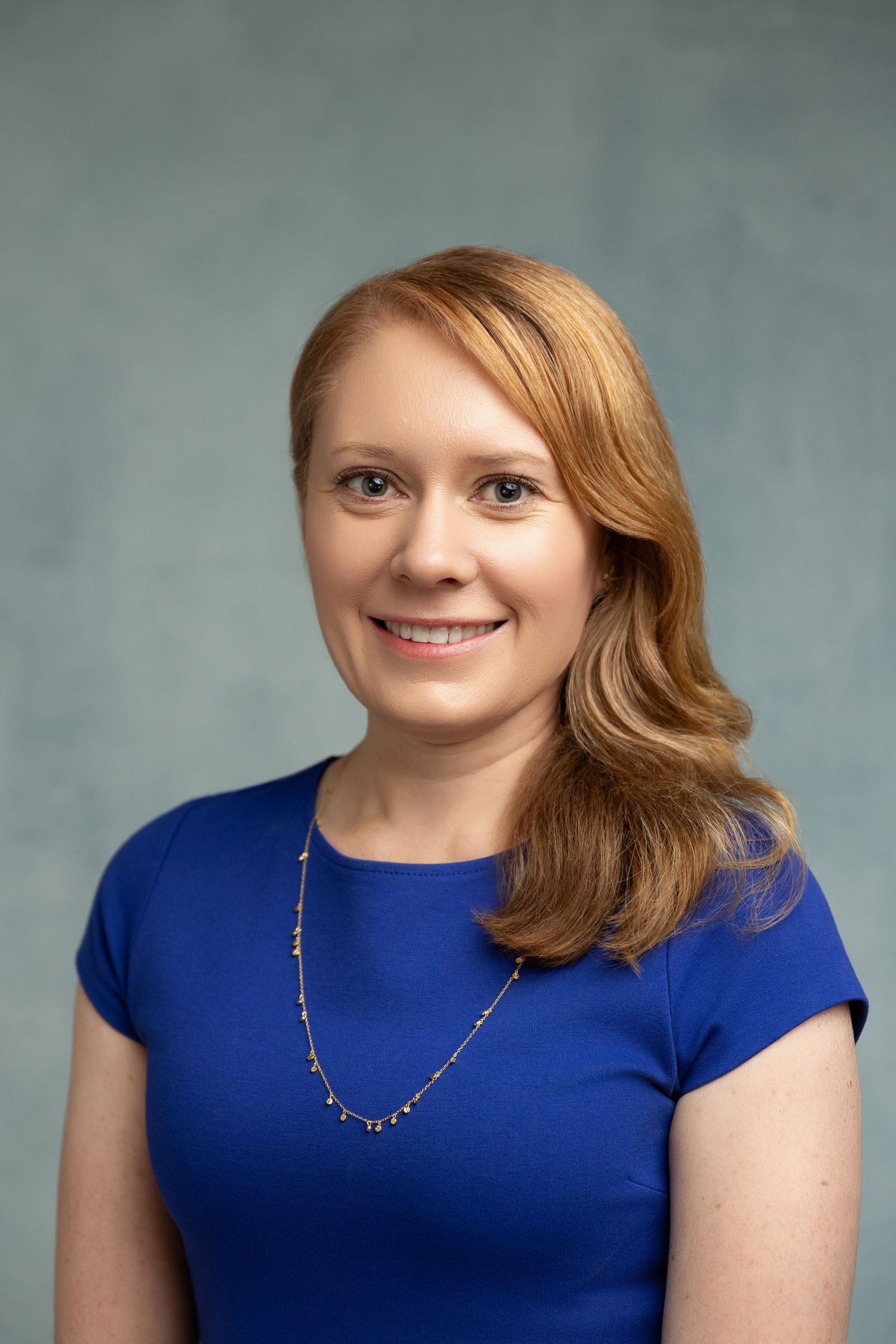 Professional headshot of a smiling woman with auburn hair wearing a blue top and gold necklace against a gray background.