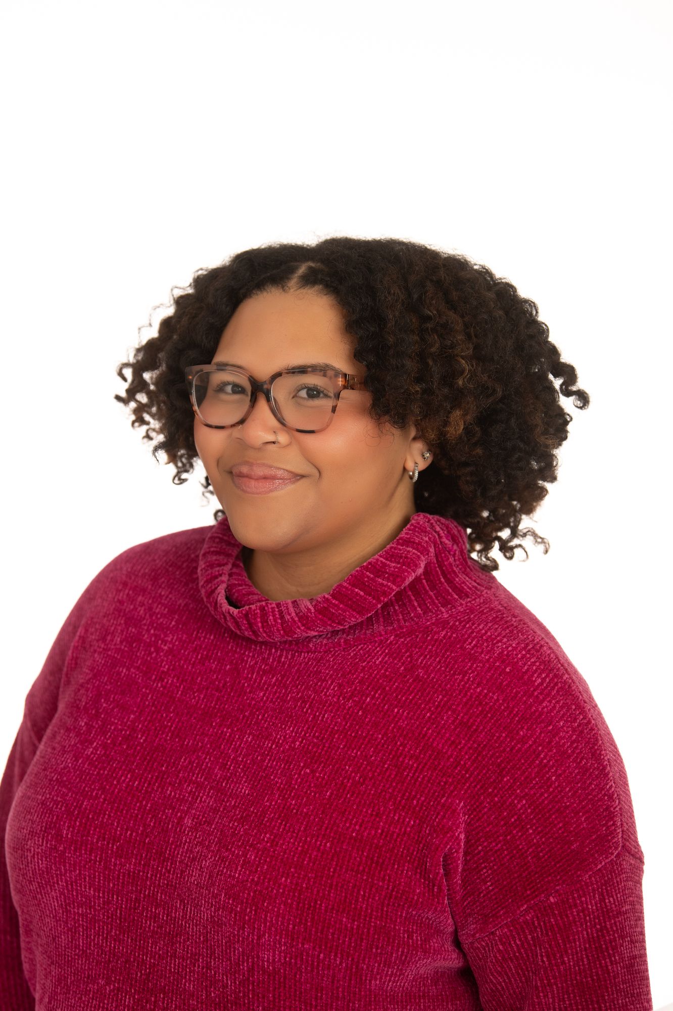 Woman with curly hair and glasses wearing a red turtleneck sweater, smiling against a white background.