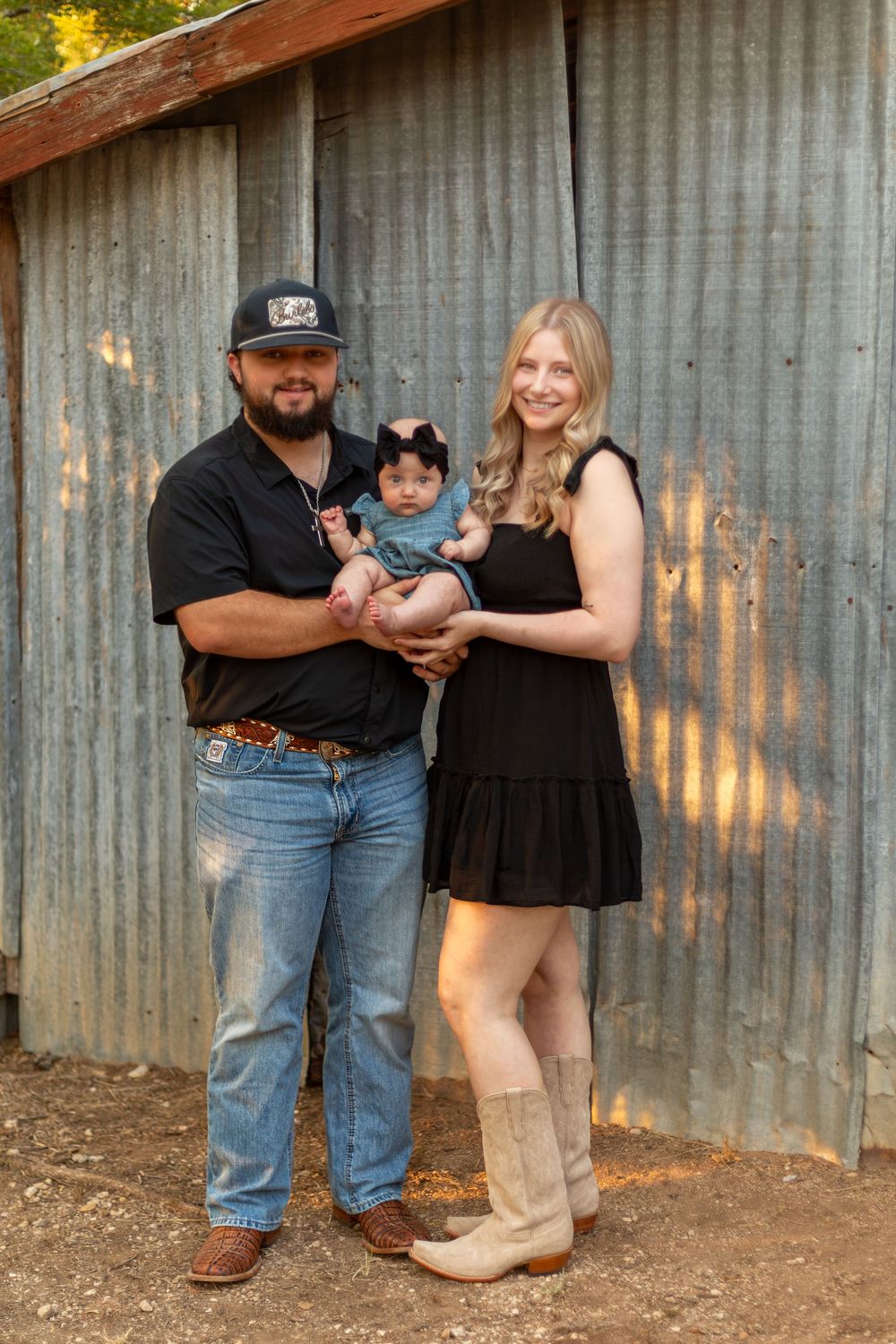 A young family poses in front of a rustic metal barn, man in black shirt and jeans holding a baby girl, woman in black dress.