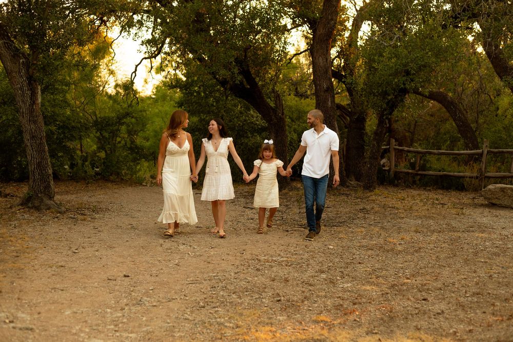 Family of four walking hand-in-hand on a dirt path through lush green trees at golden hour.