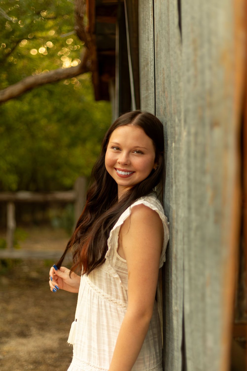 Smiling young woman with long dark hair in white dress leaning against rustic wooden wall outdoors.