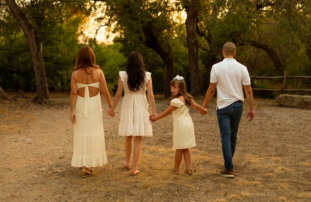 Family of four holding hands walking through a golden sunlit forest path in matching white outfits.