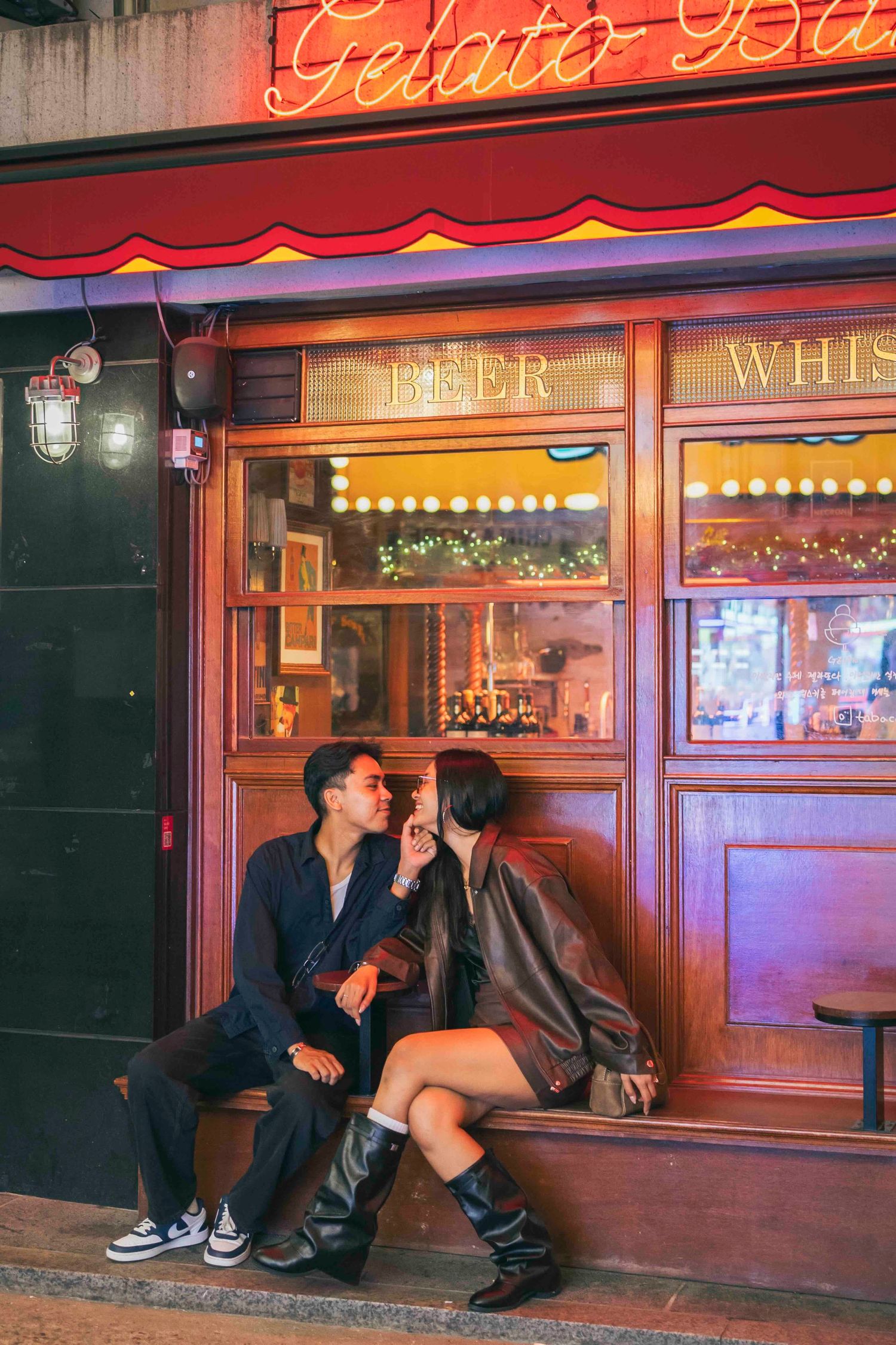 Couple sharing a romantic moment outside a cozy gelato bar with warm wooden facade and beer signs at night.