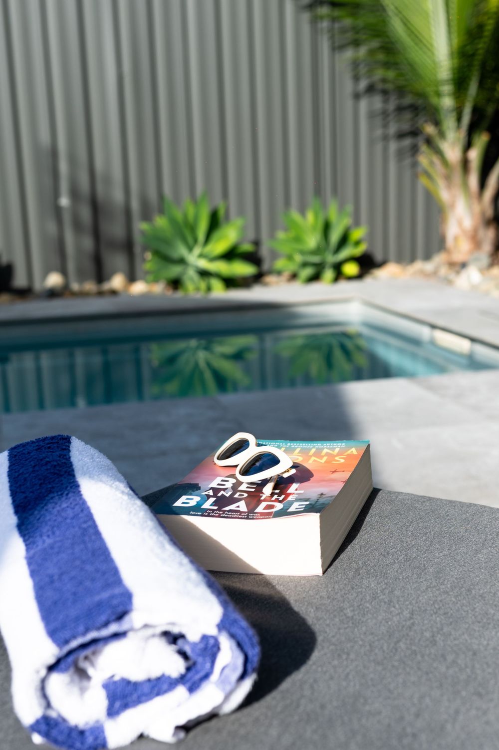 A blue striped towel and book with sunglasses rest on a pool deck beside a serene backyard swimming pool.