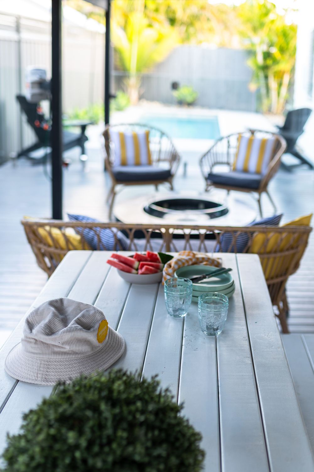 Outdoor patio table with glasses, snacks, and a hat, overlooking rattan chairs and a pool in a lush backyard.