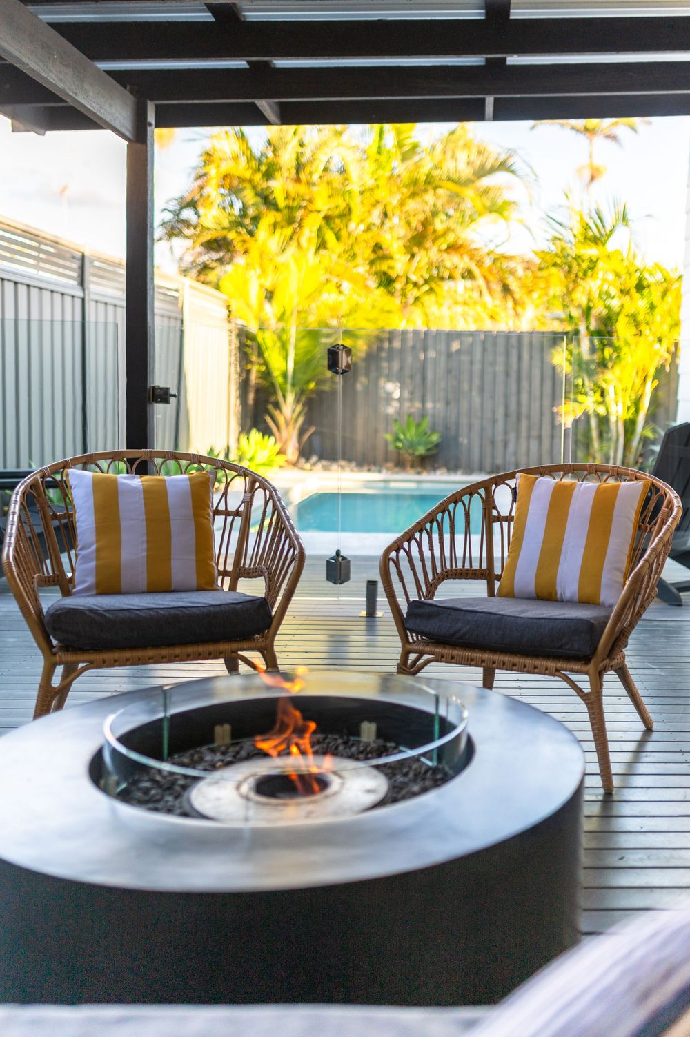 Two rattan chairs with striped cushions surround a modern fire pit on a deck beside a pool.