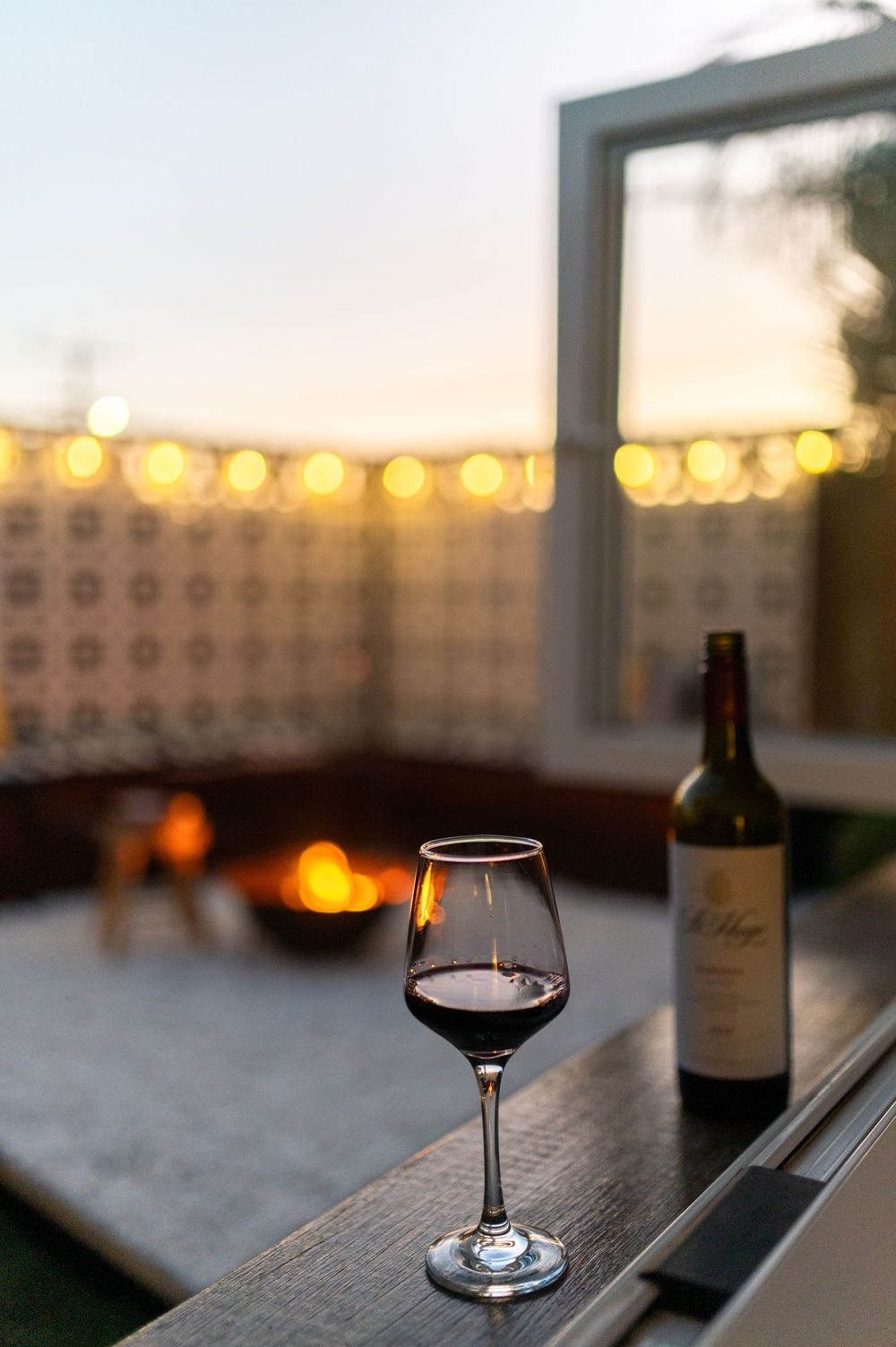 A glass of red wine on a ledge overlooking a cozy outdoor patio with string lights at sunset.