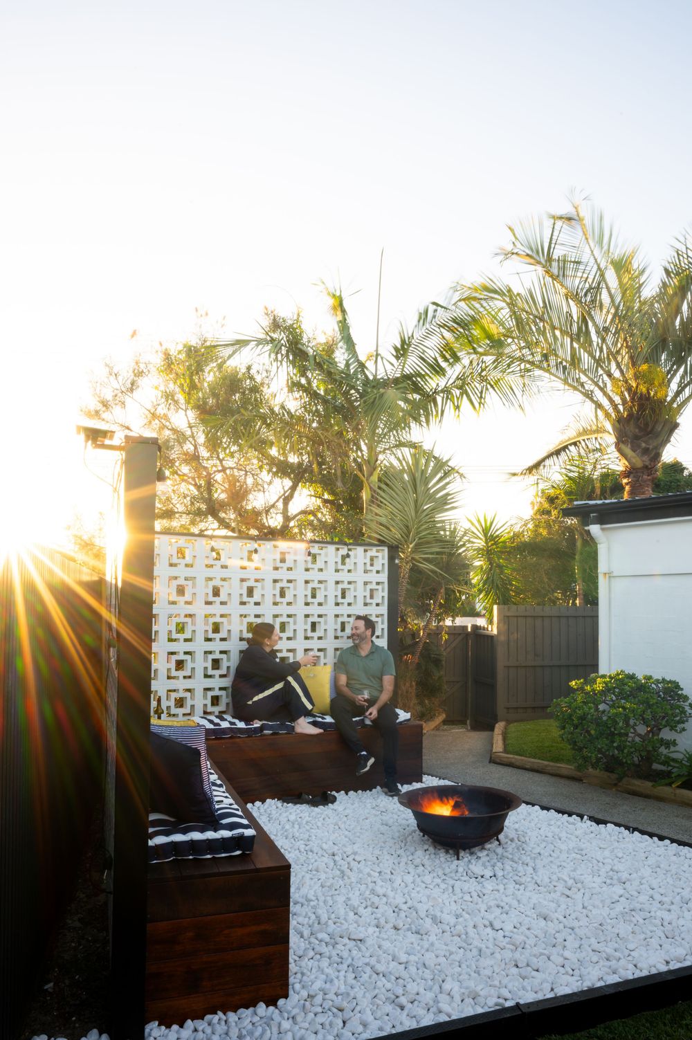 Two people sit on wooden benches in a modern outdoor patio with white gravel, a fire pit, and palm trees at sunset.