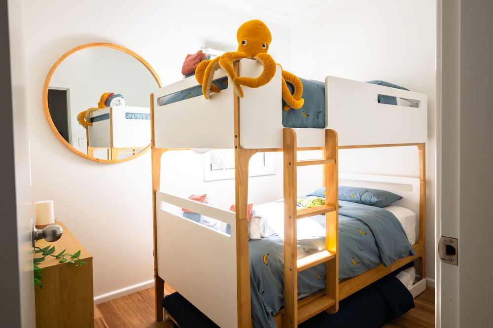 Children's wooden bunk bed with blue bedding, round mirror, and yellow octopus plush toy in a cozy kids bedroom.