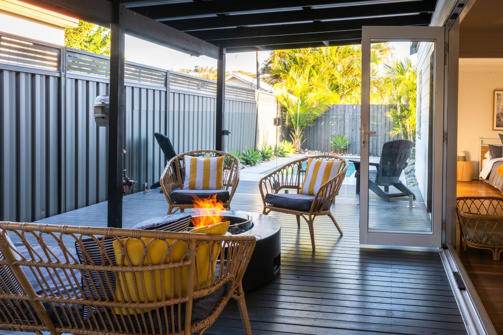 Cozy outdoor deck with rattan chairs, yellow cushions, and a fire pit, surrounded by lush greenery and modern fencing.