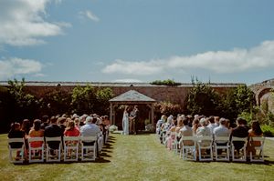 Outdoor wedding ceremony with guests seated on white chairs facing a gazebo altar in a sunny walled garden.