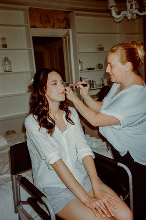 A makeup artist applies eyeshadow to a brunette bride-to-be sitting in a chair during wedding preparations.