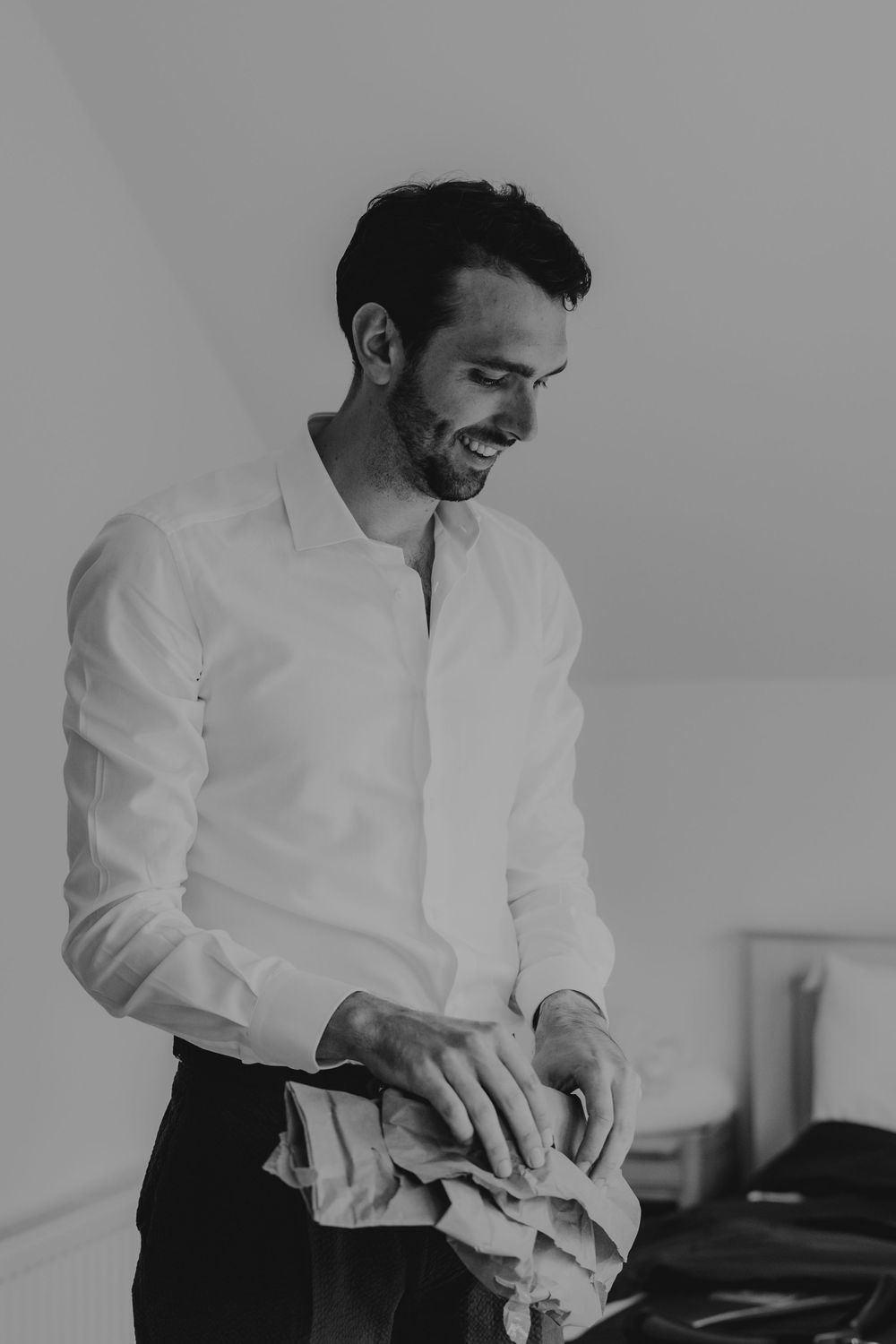 Smiling groom in white shirt preparing for his wedding day, holding clothing in a bright room.