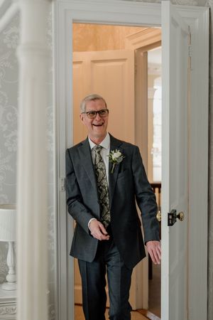 Cheerful groom in dark suit with floral tie and boutonniere entering through white double doors.