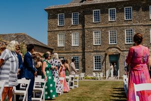 Outdoor wedding ceremony on sunny lawn beside historic stone manor house with guests seated in white chairs.