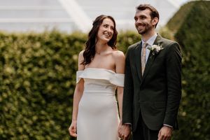 Bride in elegant off-shoulder white dress and groom in dark suit smiling outdoors at wedding.