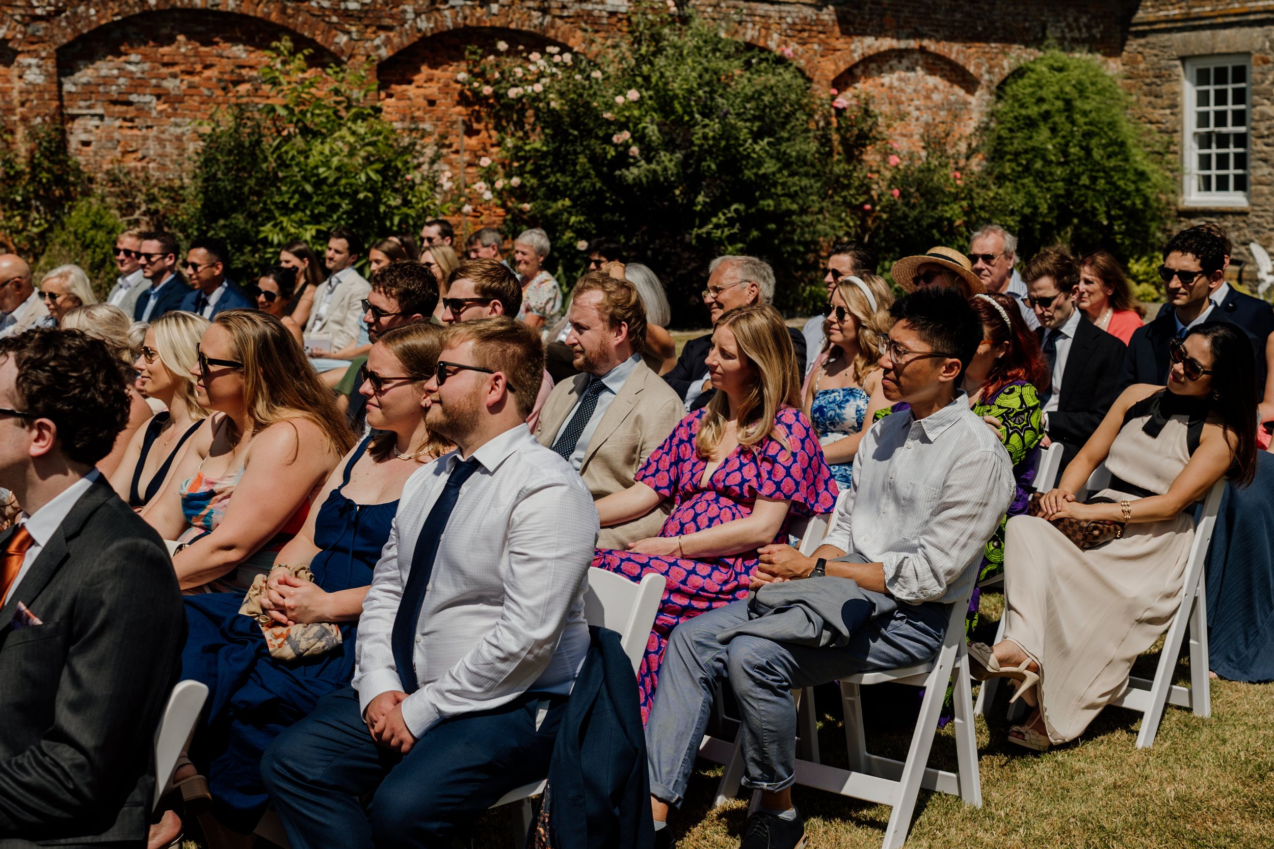 Wedding guests seated outdoors on white chairs enjoying a sunny garden ceremony at a historic brick venue.