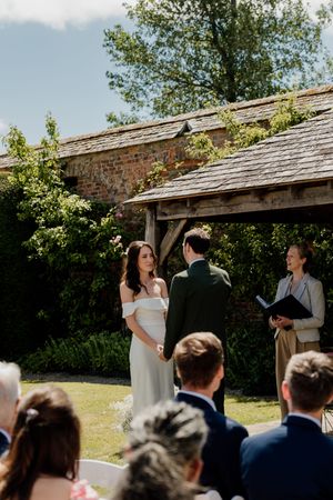 Bride and groom exchange vows outdoors at a rustic garden wedding ceremony with an officiant and seated guests.