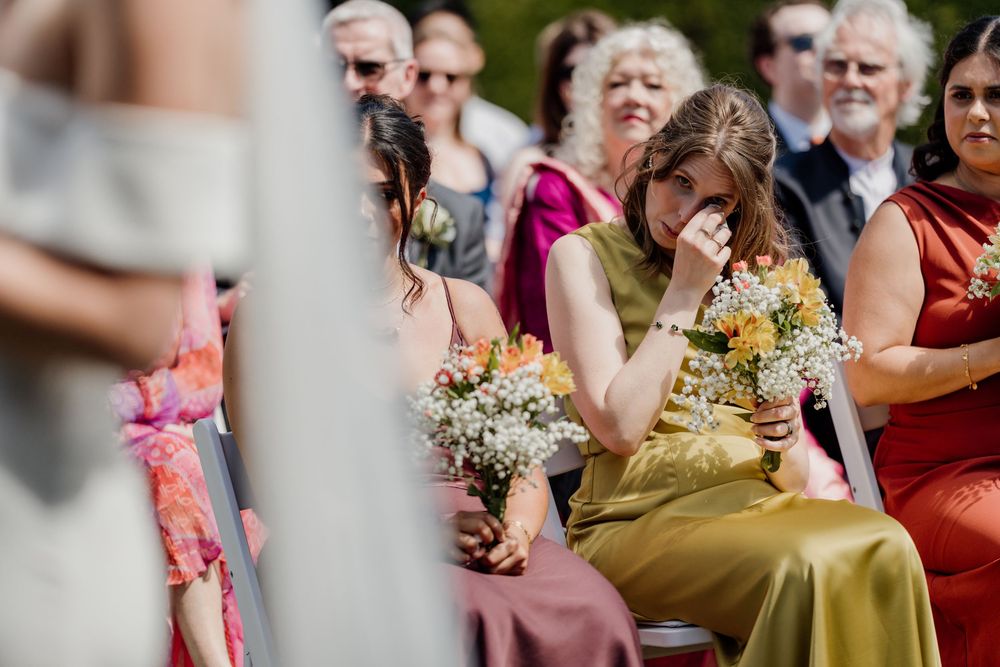 Emotional bridesmaid in olive dress wiping tears while holding bouquet during outdoor wedding ceremony.