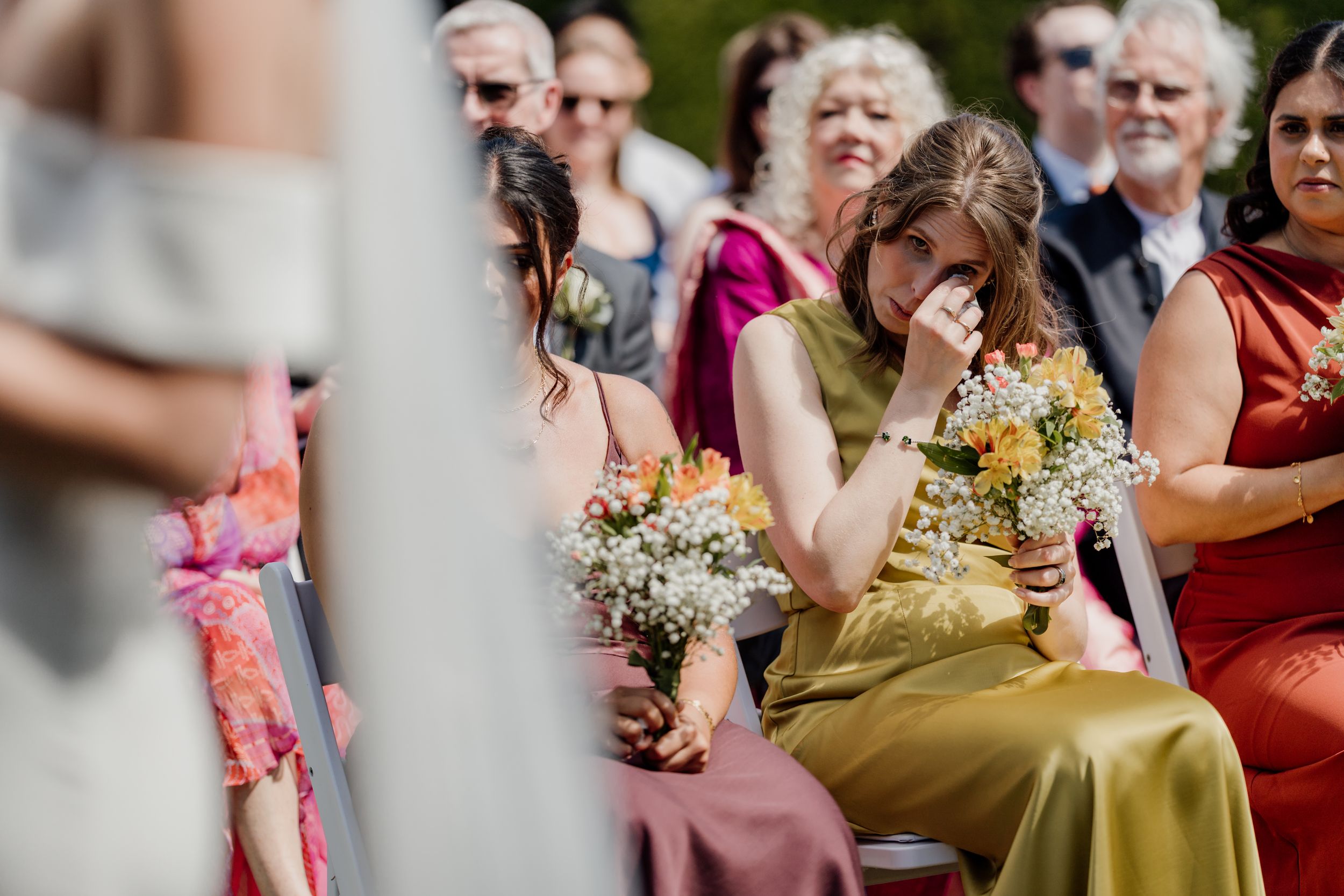 Emotional bridesmaid in olive dress wiping tears while holding bouquet during outdoor wedding ceremony.