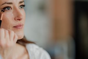 Woman applying makeup with a brush to her cheek, looking in a mirror during her beauty routine.