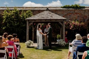 Couple exchanging vows under wooden gazebo at outdoor garden wedding ceremony with guests seated on white chairs.