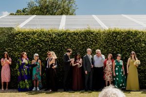 Wedding guests in colorful dresses standing in a line against a tall green hedge on a sunny day.